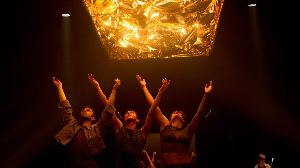 Imatge de l'espectacle, on dues noies i un noi aixequen les mans cap adalt, on hi ha un plàstic daurat il·luminat